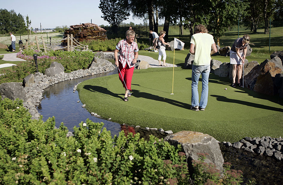 Personer spelar minigolf på grön konstgräsmatta med vattendraget och stendekorationer