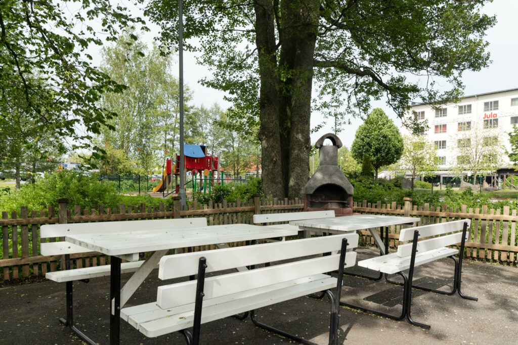 Picknickbord med bänkar vid stenugn under träd, lekplats och hotell synligt bakom