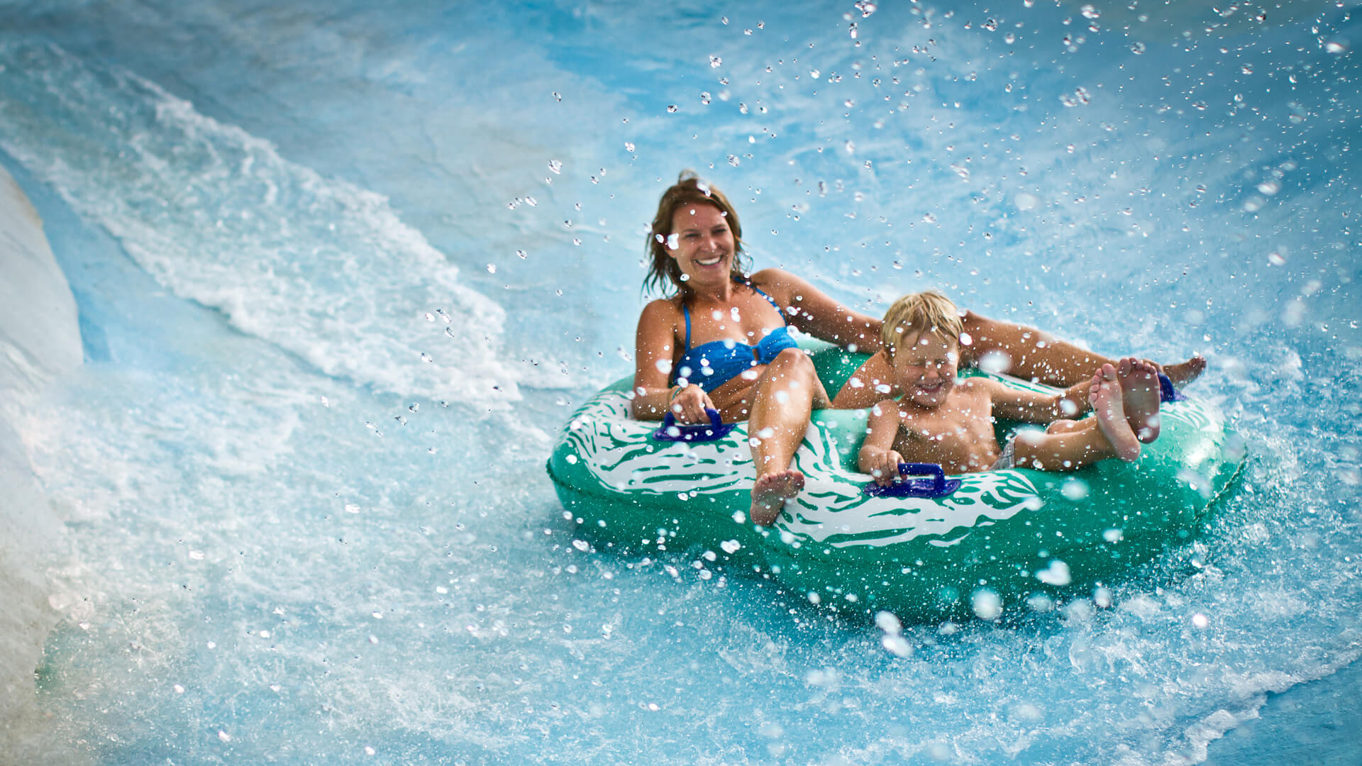 Mamma och barn åker vattenrutschkana på grön badring med vattenstänk