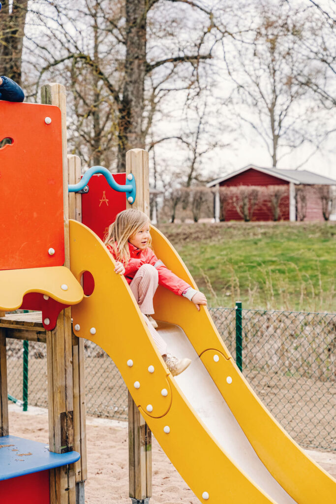 Liten flicka i röd jacka åker rutschkana på lekplats med träd i bakgrunden