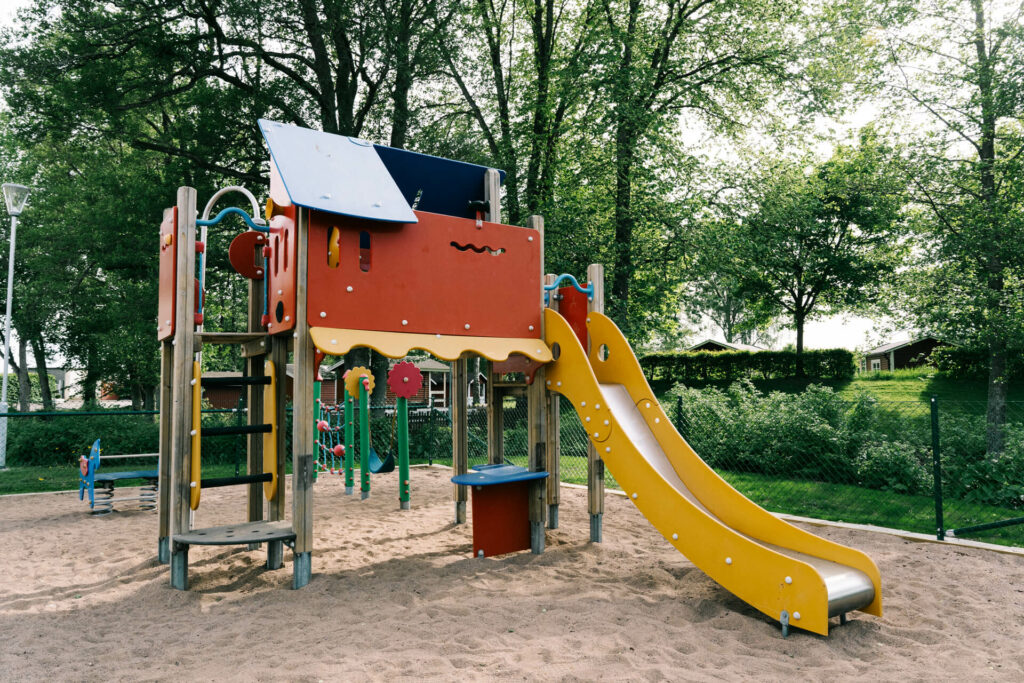 Färgglad lekplats med klätterställning, gul rutschkana och sandunderlag omgiven av träd