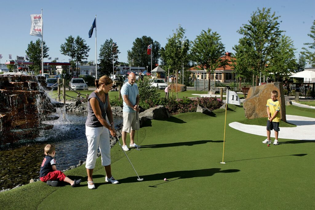 Familj med barn spelar minigolf vid fontän och konstgjord å i parkliknande miljö