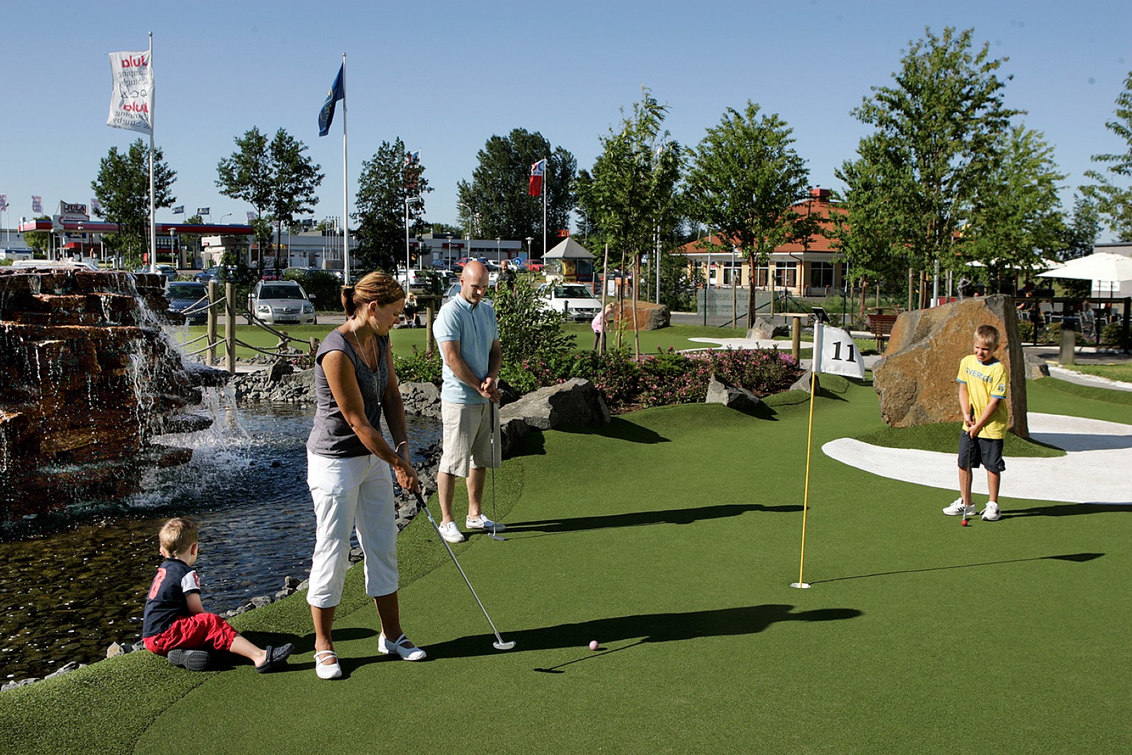 Familj med barn spelar minigolf vid fontän och konstgjord å i parkliknande miljö
